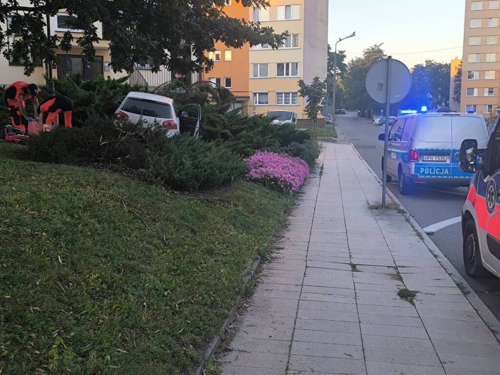 Ratownicy medyczni udzielają pomocy osobie w rozbitym samochodzie obok policyjnego radiowozu na osiedlowej ulicy.