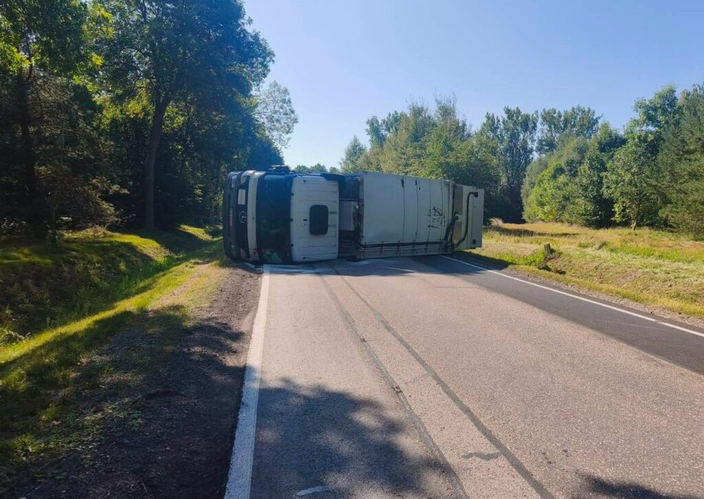 Ciągnik siodłowy leży na boku na poboczu drogi, otoczony przez zieleń drzew i krzewów.