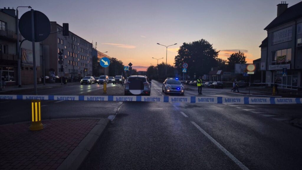Policyjny radiowóz stoi za barierką zabezpieczającą, a funkcjonariusz kieruje ruchem na zablokowanej ulicy o zmierzchu.
