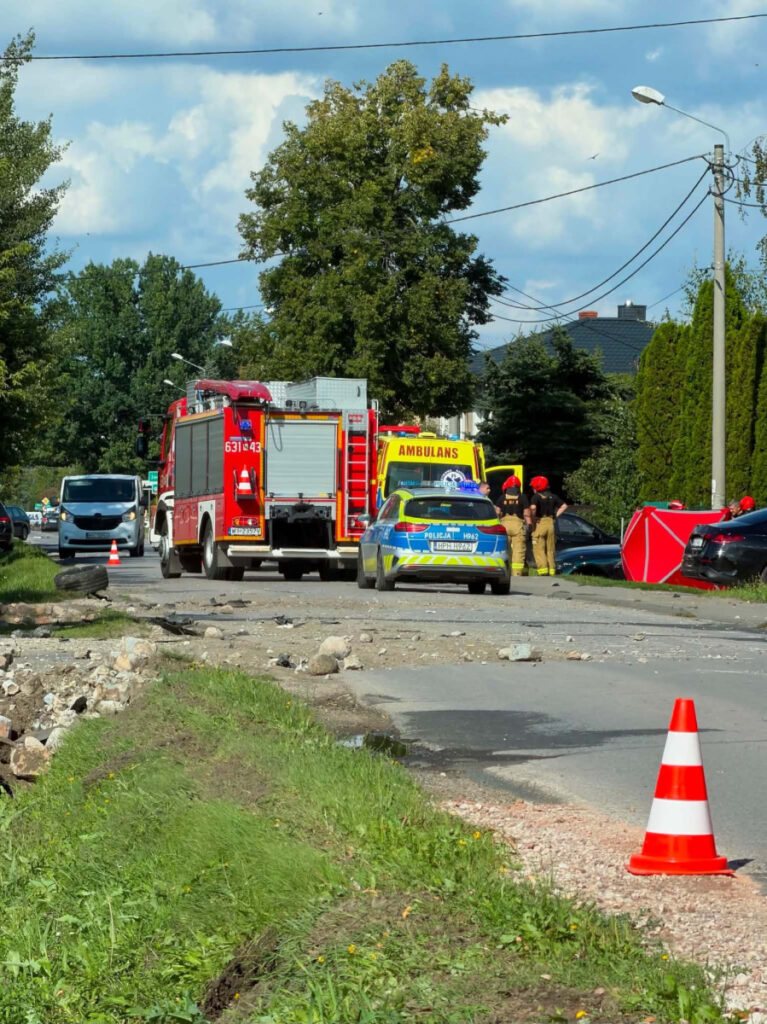 W tle widać straż pożarną i karetkę, a wokół grupę ratowników pracujących przy uszkodzonym pojeździe.