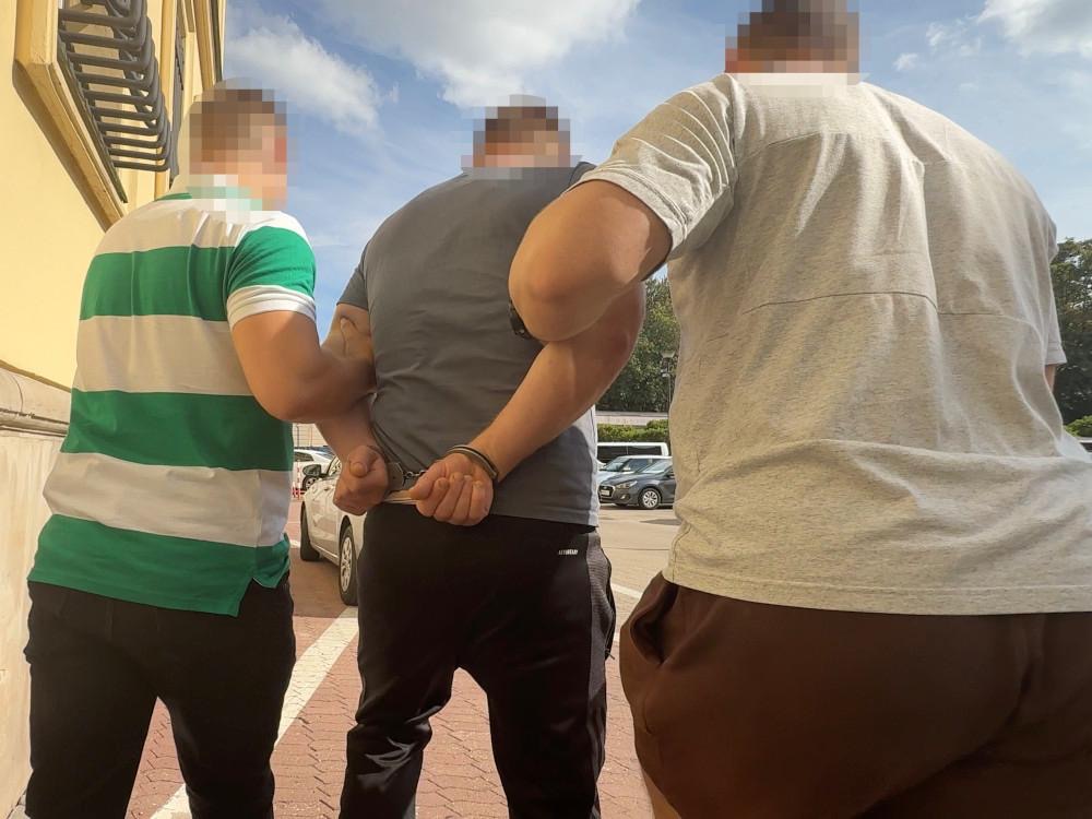 Mężczyzna w kajdankach prowadzony przez dwóch mężczyzn na ulicy, w tle zaparkowane samochody.