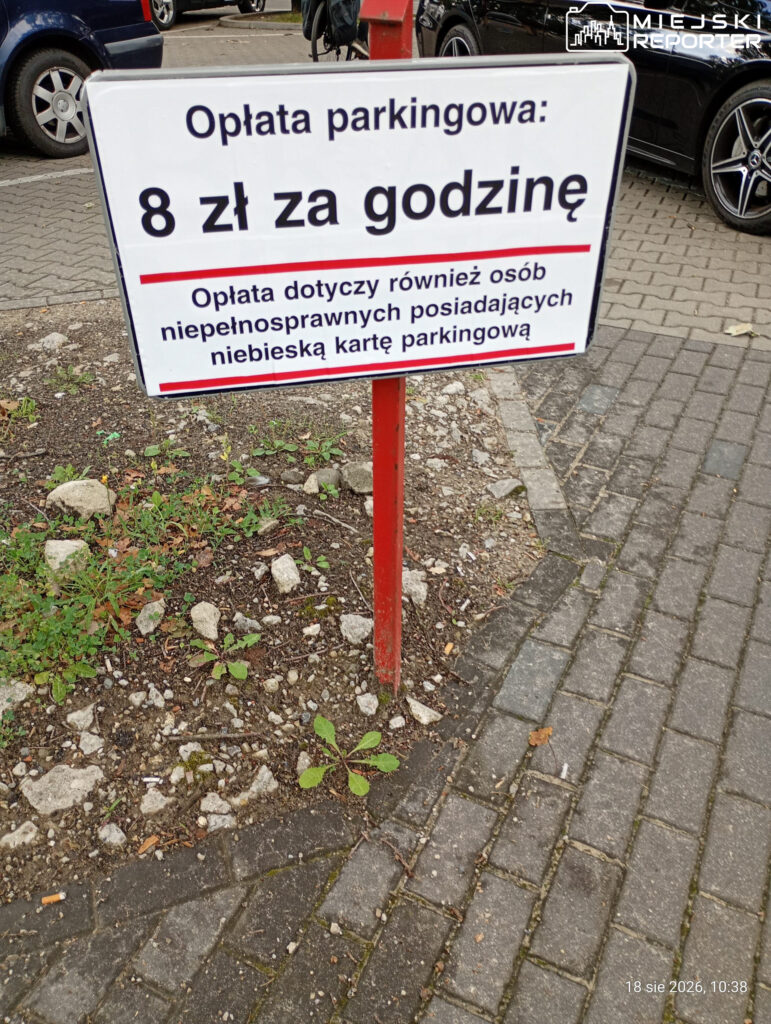 Tablica informacyjna z napisem "Opłata parkingowa: 8 zł za godzinę" w otoczeniu parkingu, z widocznymi roślinami i kostką brukową.
