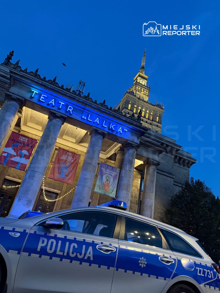 Policyjny radiowóz zaparkowany przed Teatrem "Lalka", oświetlonym niebieskimi światłami w wieczornym otoczeniu.
