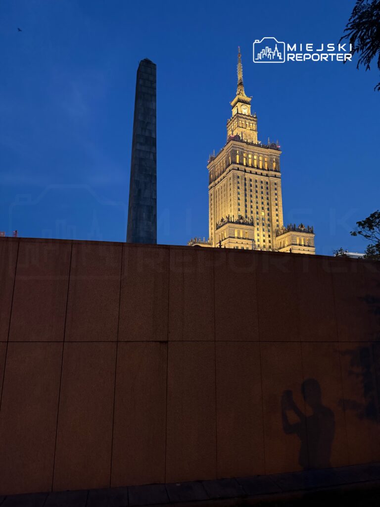Wysoki budynek oświetlony nocą, z obeliskiem w tle, a w cieniu widoczna sylwetka osoby trzymającej telefon.