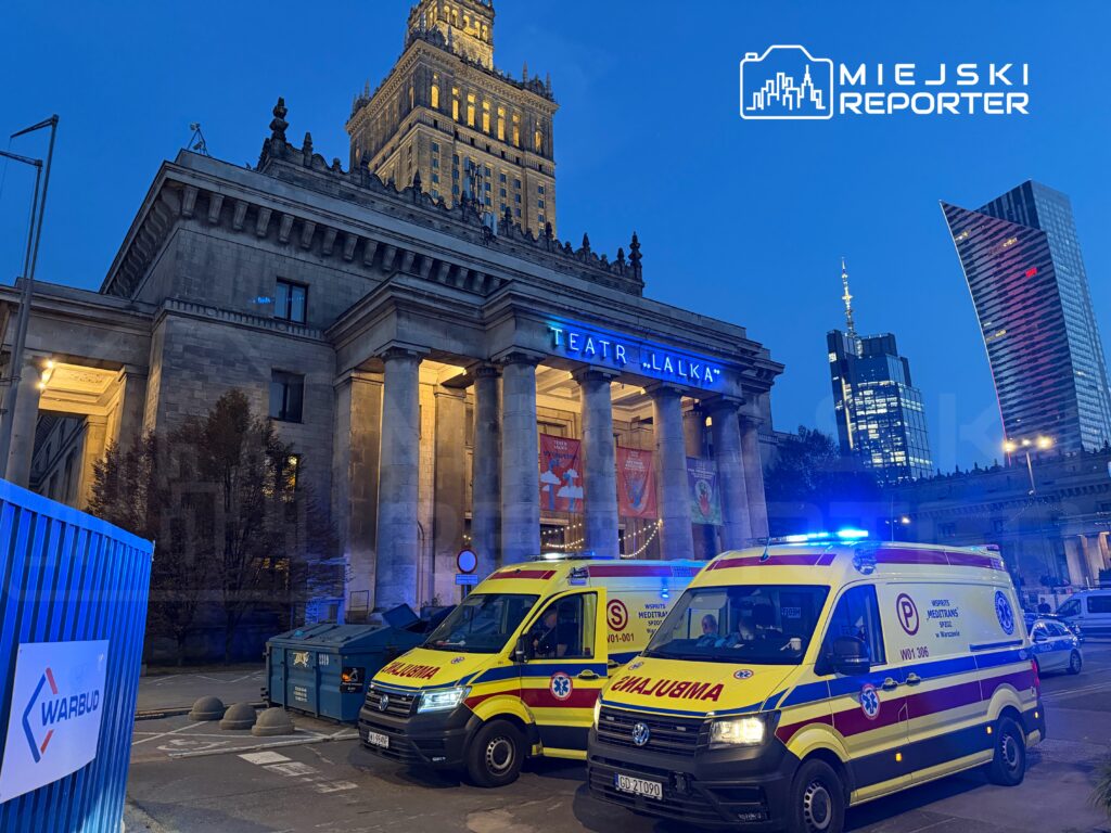 Dwa karetki pogotowia z włączonymi sygnałami świetlnymi stoją przed Teatrem "Lalka" w Warszawie, w otoczeniu nowoczesnych budynków.
