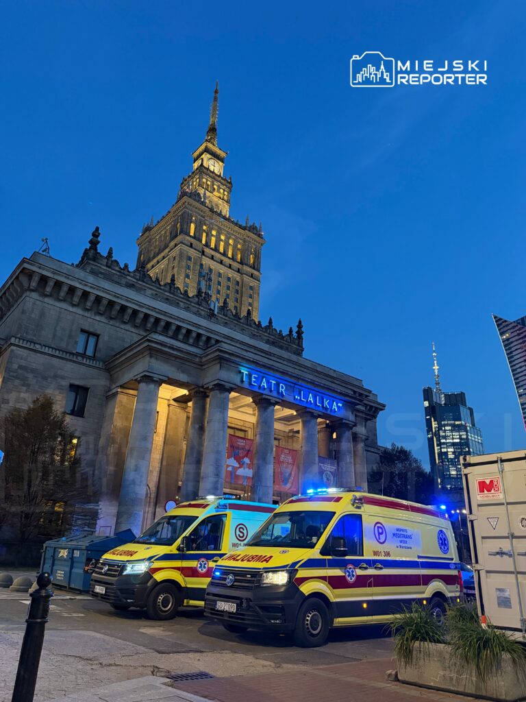 Dwa ambulansy z włączonymi sygnałami świetlnymi stoją przed Teatrem "Lalka" w wieczornym oświetleniu.