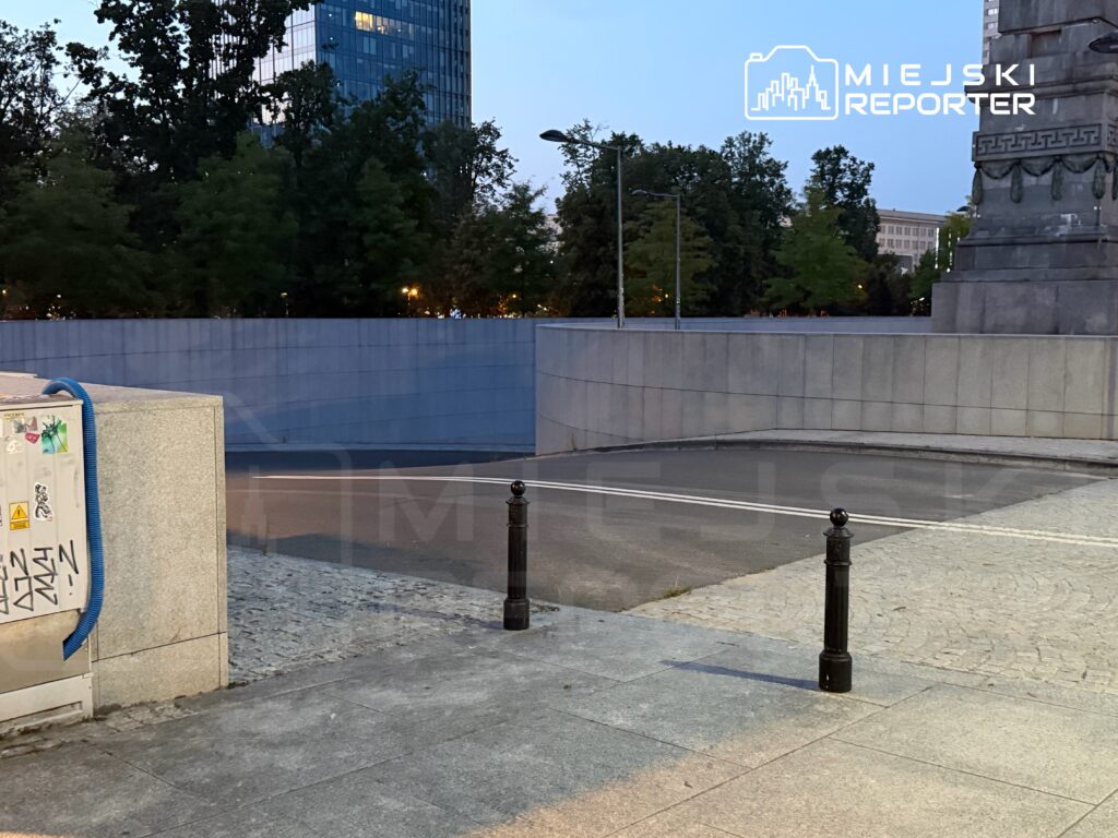 Pusty plac z granitową nawierzchnią, otoczony drzewami i nowoczesnym biurowcem, w tle widoczny monument.