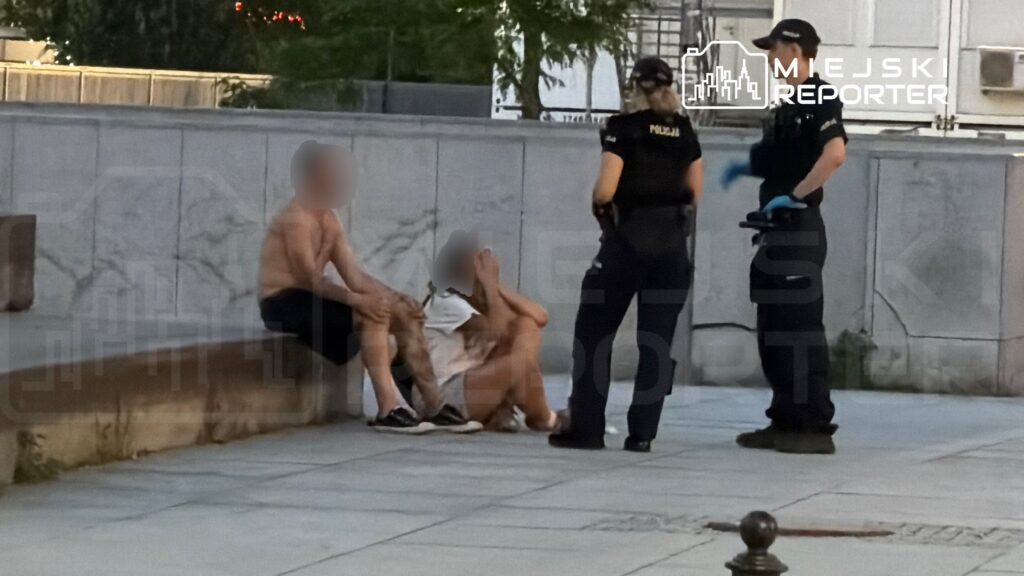 Dwóch mężczyzn siedzi na ławce, a obok nich stoją policjanci rozmawiający z nimi w miejskim otoczeniu.
