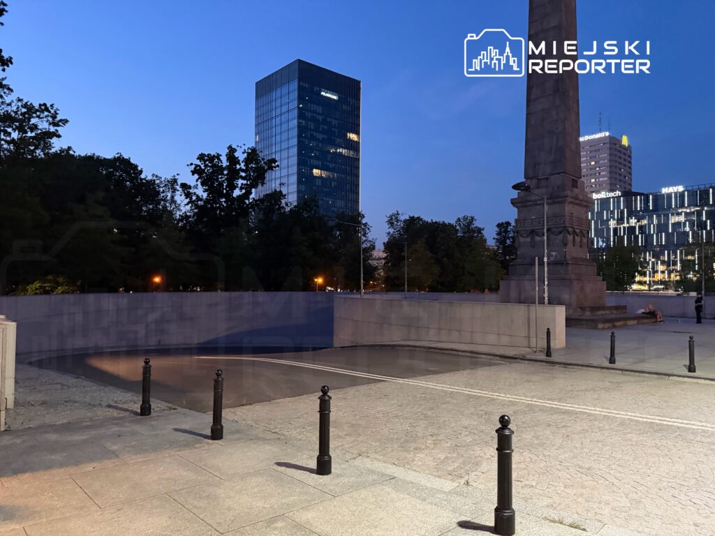 Nowoczesny biurowiec z przeszkloną fasadą w tle, obelisk w centralnej części, oświetlona przestrzeń publiczna wieczorem.