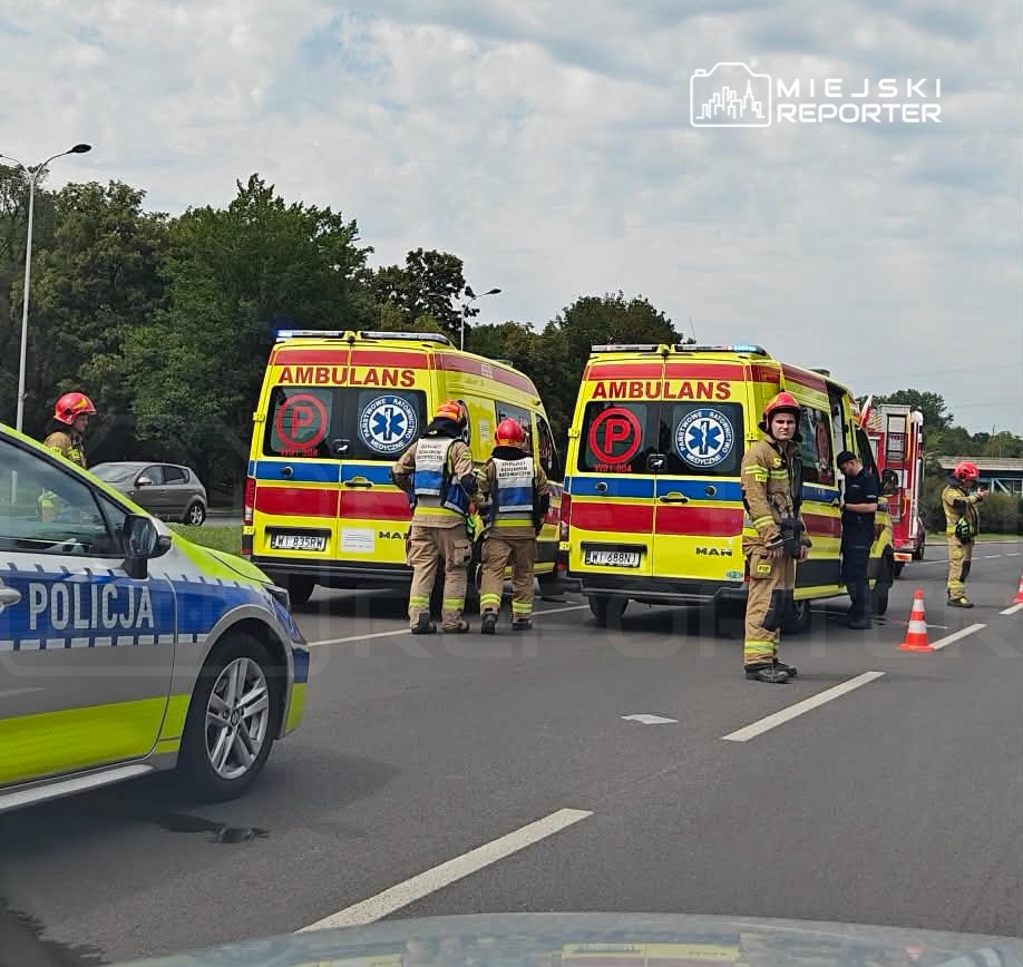 Grupa strażaków i ratowników medycznych pracuje przy dwóch ambulansach na drodze, w otoczeniu drzew.