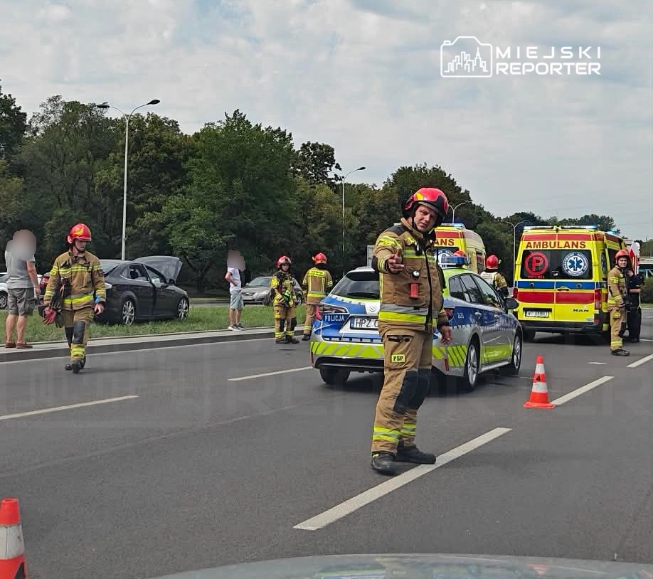 Strażacy w hełmach kierują ruchem na drodze, obok stoją pojazdy służb ratunkowych.