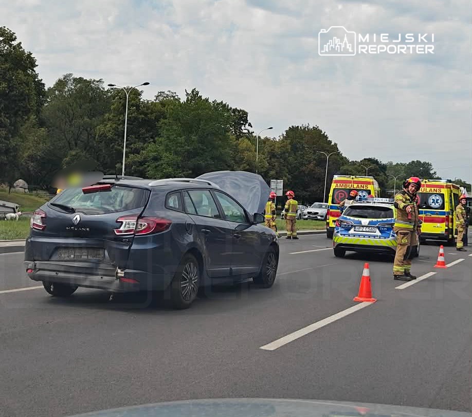 Na drodze stoi niebieski samochód osobowy z otwartą maską, obok pracują strażacy w hełmach, a w tle widać karetki pogotowia.