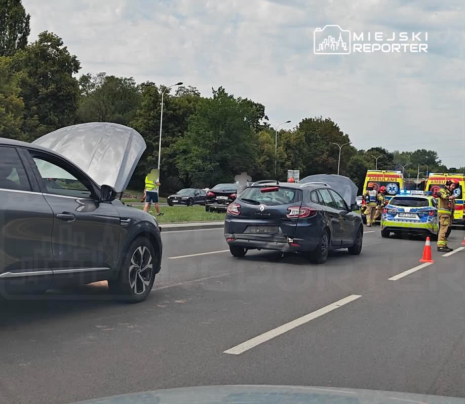 Czarny samochód z otwartą maską stoi na drodze, a obok niego zespół ratunkowy udziela pomocy.