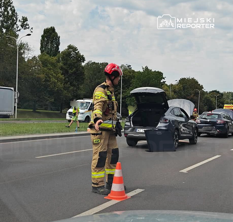 Strażak w hełmie kieruje ruchem na drodze, obok otwartego samochodu z podniesioną maską.
