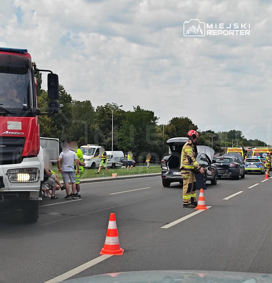 Grupa ratowników w kamizelkach odblaskowych udziela pomocy poszkodowanemu na drodze, obok stoi wóz strażacki.