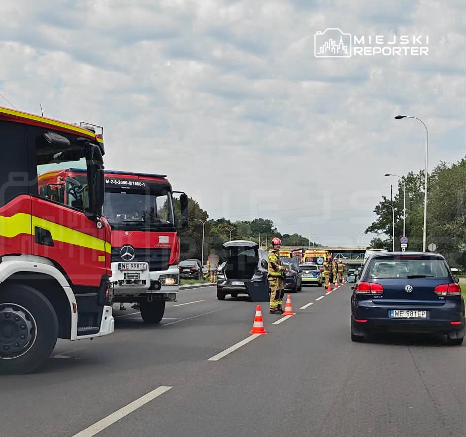 Na drodze stoją dwa wozy strażackie, a strażacy w mundurach rozmawiają z kierowcą samochodu osobowego.