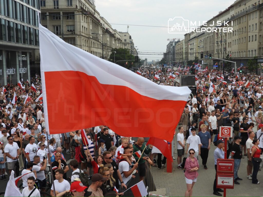 Tłum ludzi trzymający flagi, w tym polską, podczas manifestacji na ulicy w miejskim otoczeniu.
