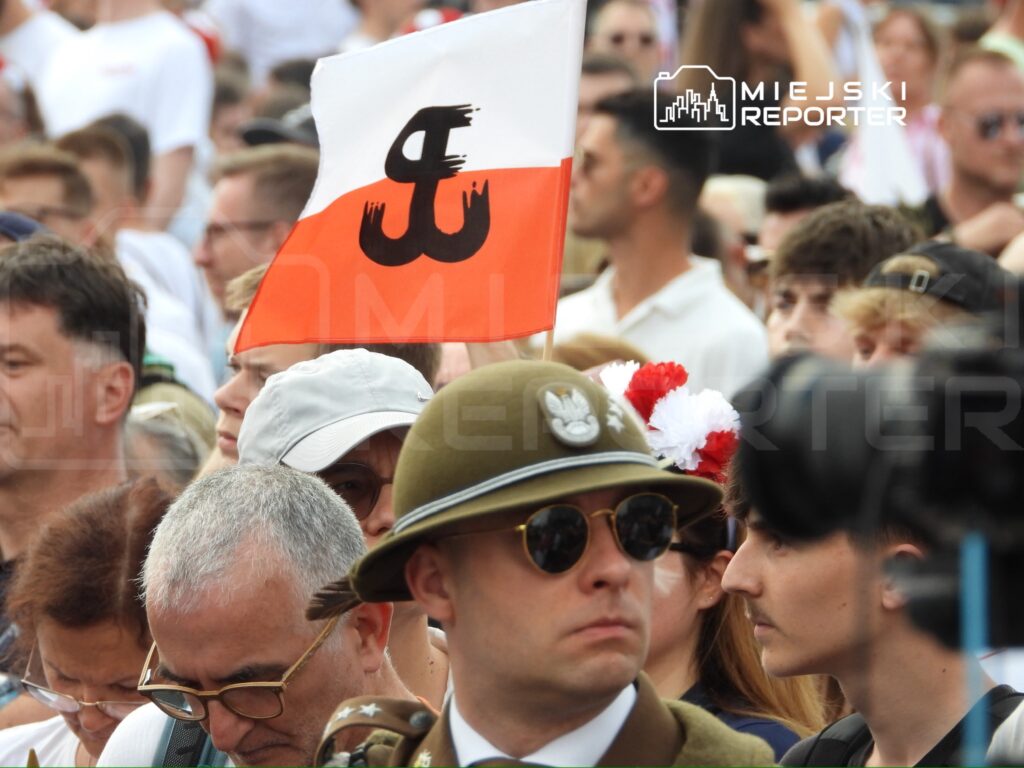 Grupa ludzi na zgromadzeniu, w tle uniesiona flaga z symbolem "kotwicy" w barwach biało-czerwonych.