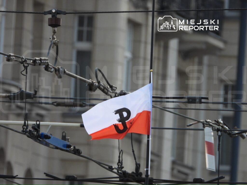 Flaga z symbolem "JP" powiewa na tle ulicznych przewodów elektrycznych w miejskiej scenerii.