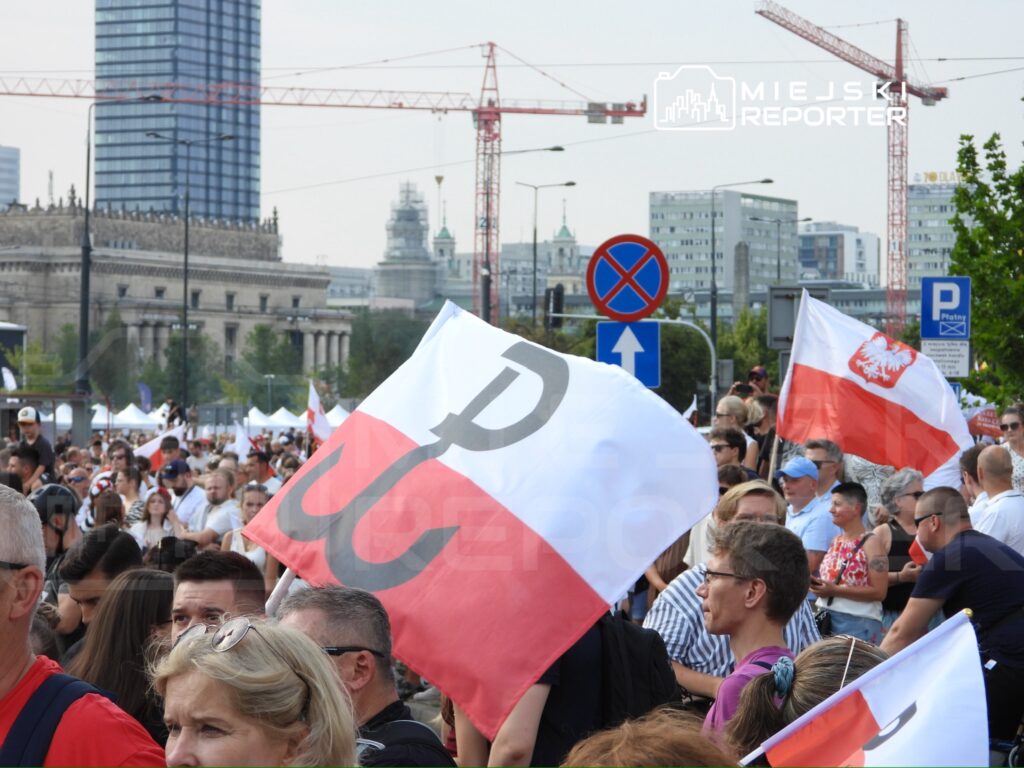 Tłum ludzi z flagami w rękach uczestniczy w wydarzeniu na ulicy, z nowoczesnymi budynkami w tle.