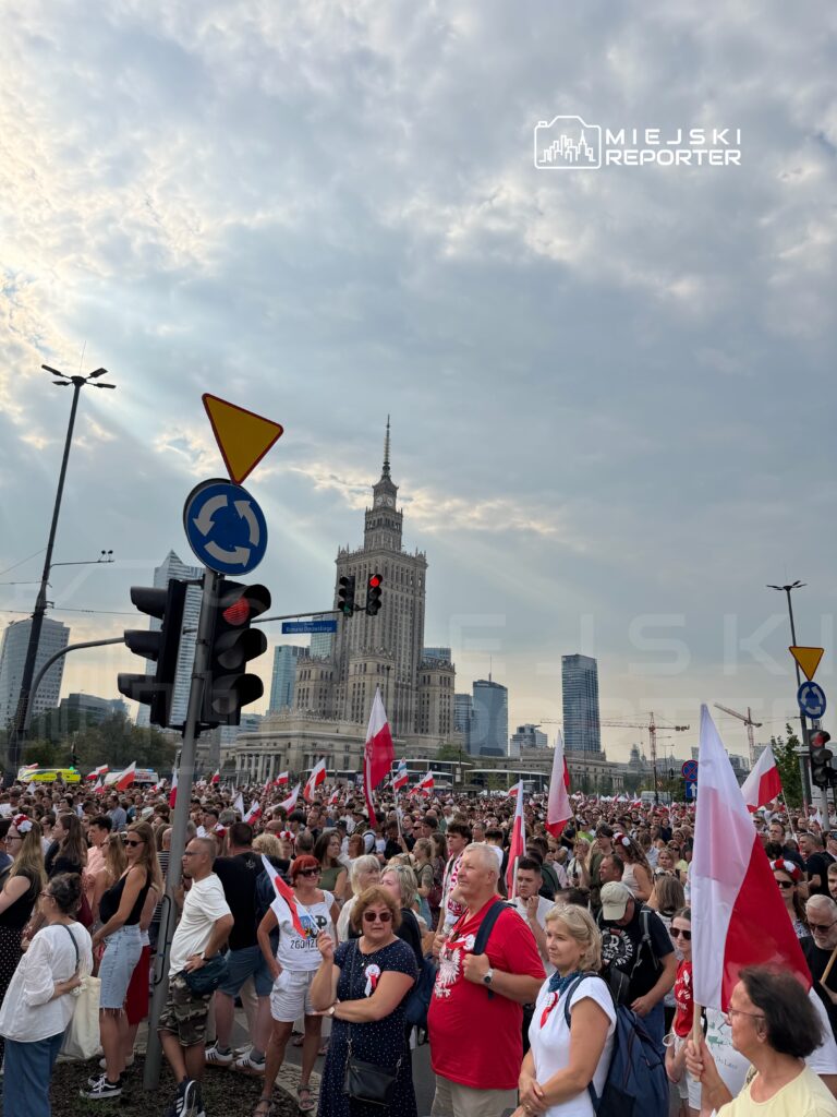 Tłum ludzi z flagami Polski gromadzi się na placu przed nowoczesnym biurowcem, w tle widoczny Pałac Kultury i Nauki.
