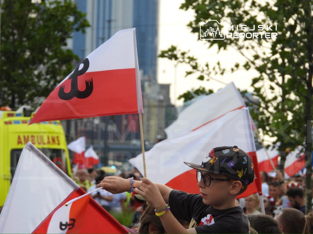 Chłopiec w czapce trzyma flagę z symbolem Polski, otoczony przez innych uczestników manifestacji.