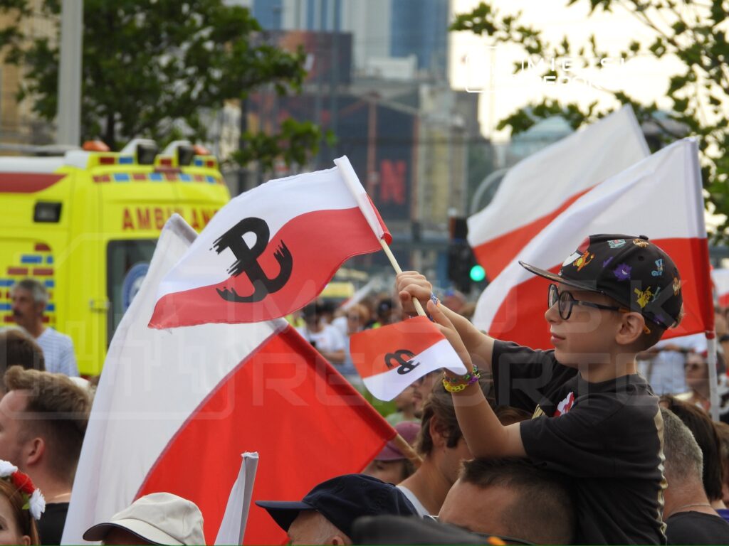 Chłopiec w okularach trzyma flagi z symbolem Polski Walczącej, siedząc na ramionach dorosłego w tłumie.