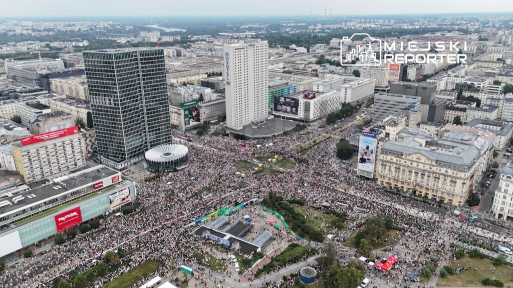 Tłum ludzi uczestniczy w demonstracji na miejskim placu, otoczonym nowoczesnymi budynkami i zielenią.
