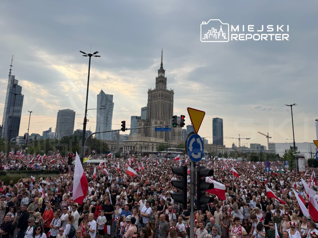 Tłum ludzi z flagami w rękach uczestniczy w demonstracji na ulicy, w tle widoczny Pałac Kultury i Nauki.
