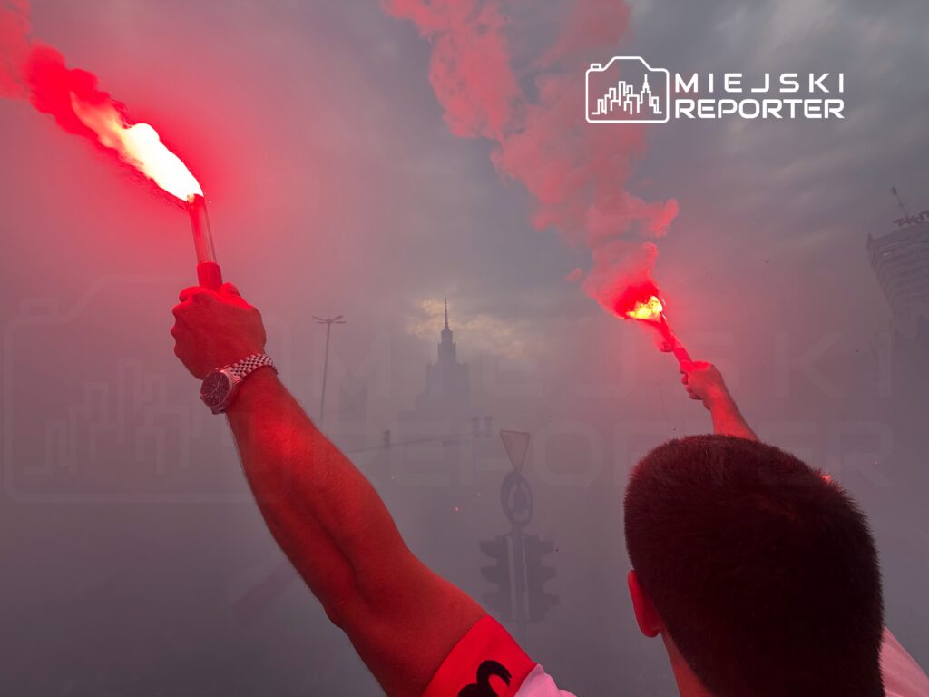 Mężczyzna trzymający czerwoną racę w gęstej mgle, w tle widoczny zarys miejskich budynków.