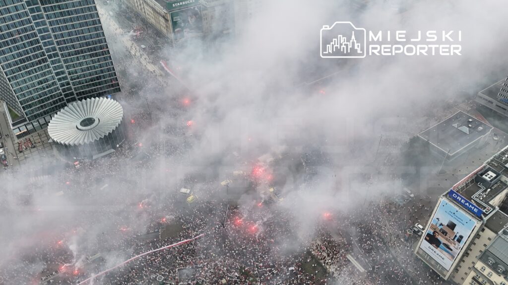 Tłum ludzi z flagami narodowymi gromadzi się w centrum miasta, otoczony gęstym dymem i fajerwerkami.