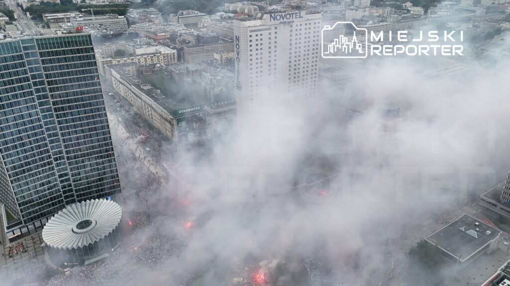 Tłum ludzi w dymie z fajerwerków gromadzi się na ulicy, otoczony nowoczesnymi budynkami.