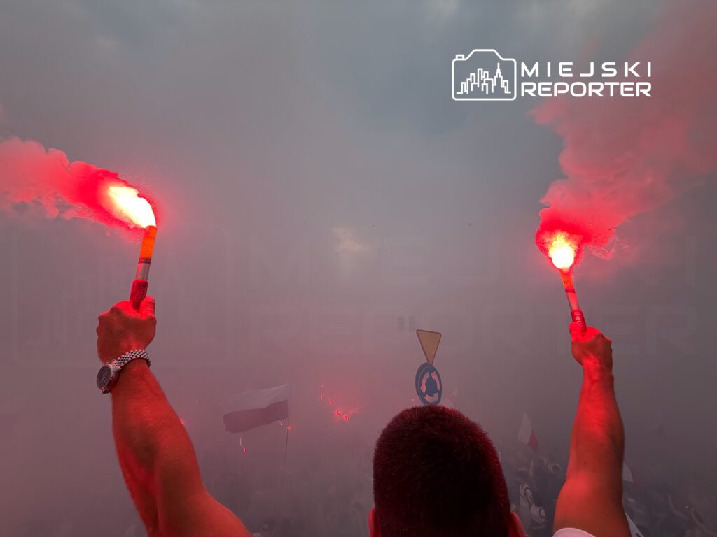 Osoba trzymająca racę świetlną w tłumie, otoczona gęstym dymem, z widocznymi flagami w tle.