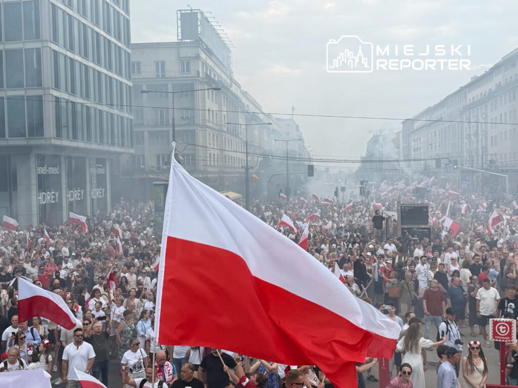 Tłum ludzi z flagami w dłoniach uczestniczy w manifestacji na ulicy, w tle widoczne budynki i lekki dym.