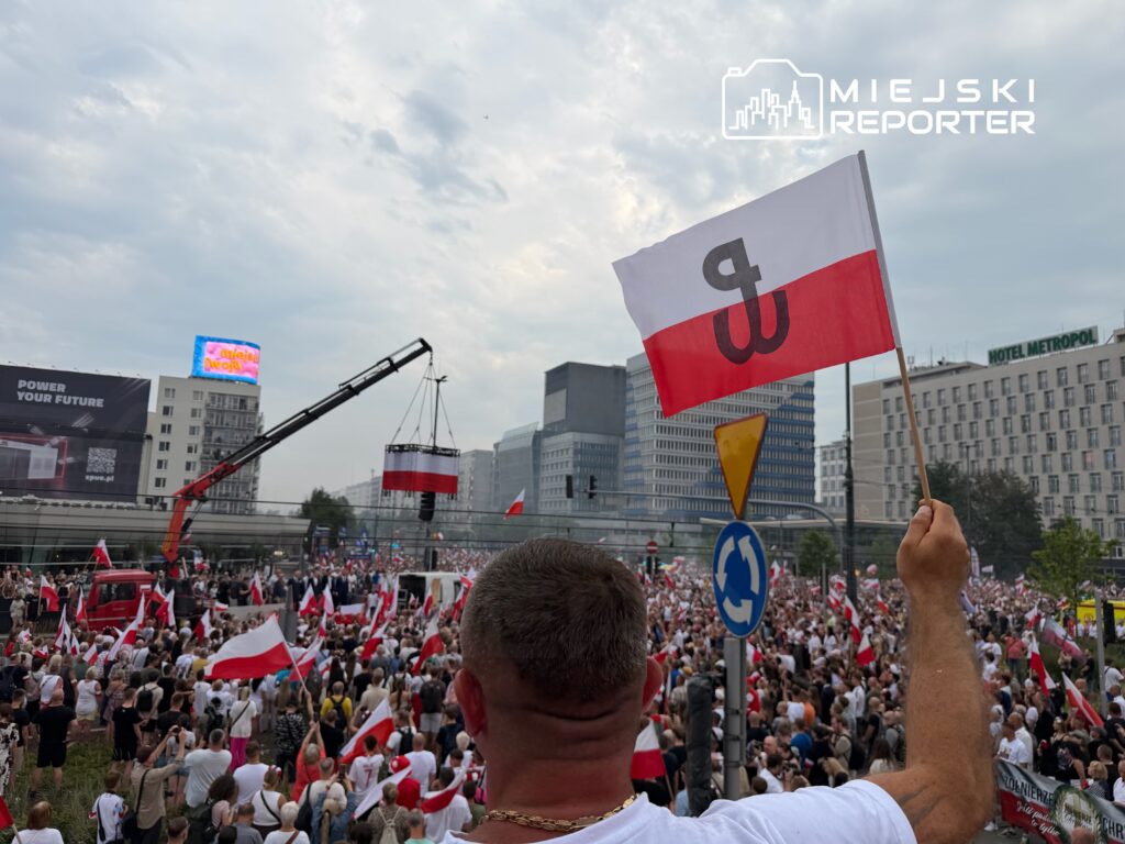 Tłum ludzi z flagami w rękach gromadzi się na ulicy, a w tle dźwiga podnosi transparent z symbolem.