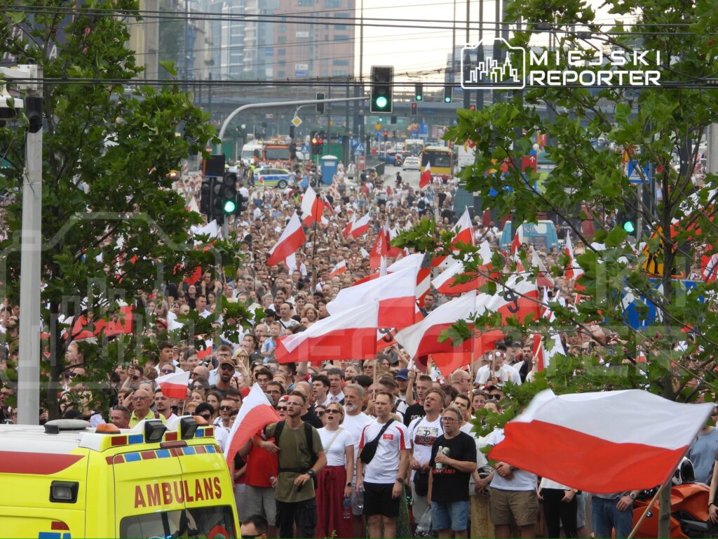 Tłum ludzi z biało-czerwonymi flagami maszeruje wzdłuż ulicy, otoczeni nowoczesnymi budynkami.