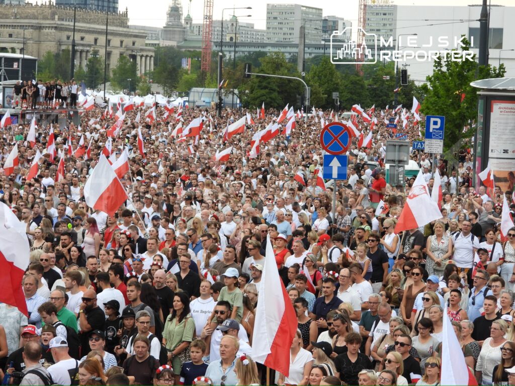 Tłum ludzi trzymających biało-czerwone flagi, zgromadzony na ulicy w centrum miasta podczas wydarzenia publicznego.