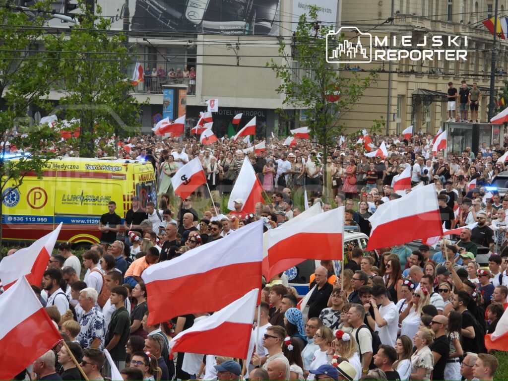 Tłum ludzi z biało-czerwonymi flagami uczestniczy w demonstracji na ulicach miasta. W tle widać ambulans.