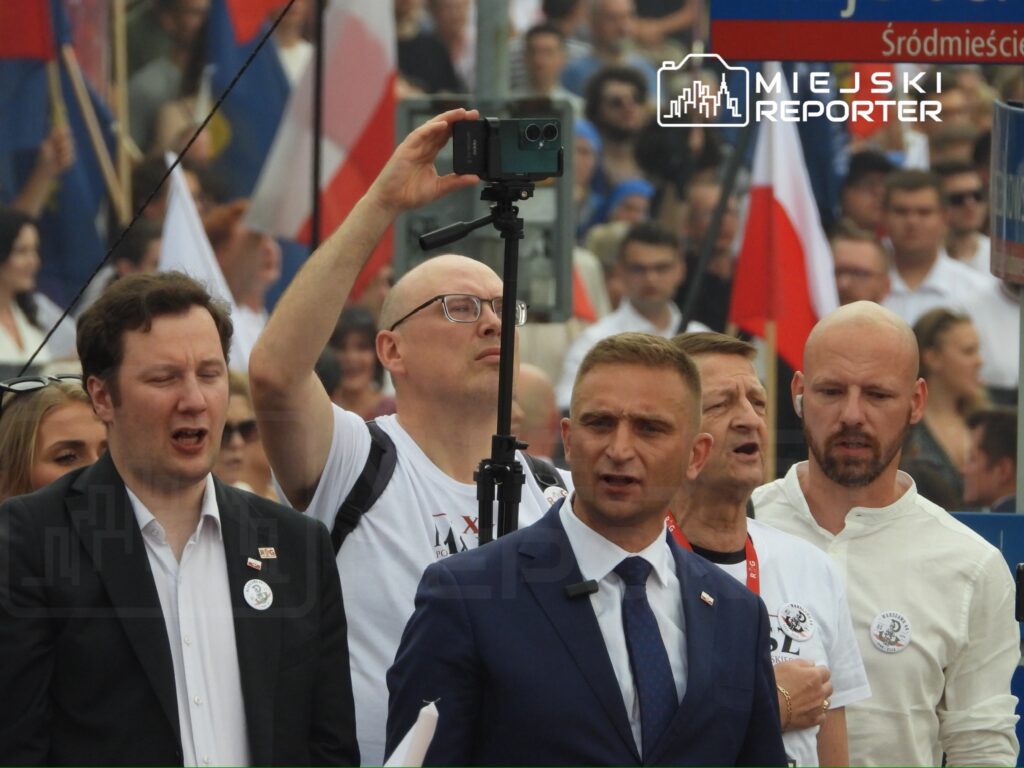 Grupa mężczyzn w garniturach i koszulach białych śpiewa na demonstracji, trzymając flagi i transparenty.