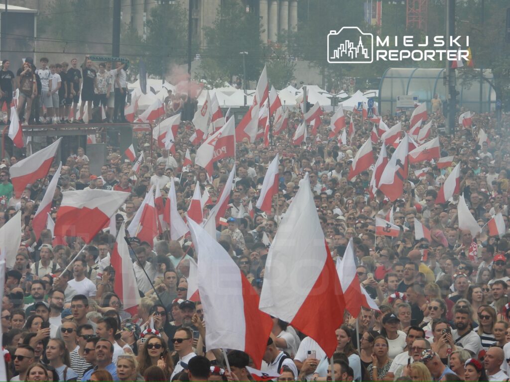 Tłum ludzi trzymających polskie flagi, gromadzący się na wydarzeniu na otwartej przestrzeni.