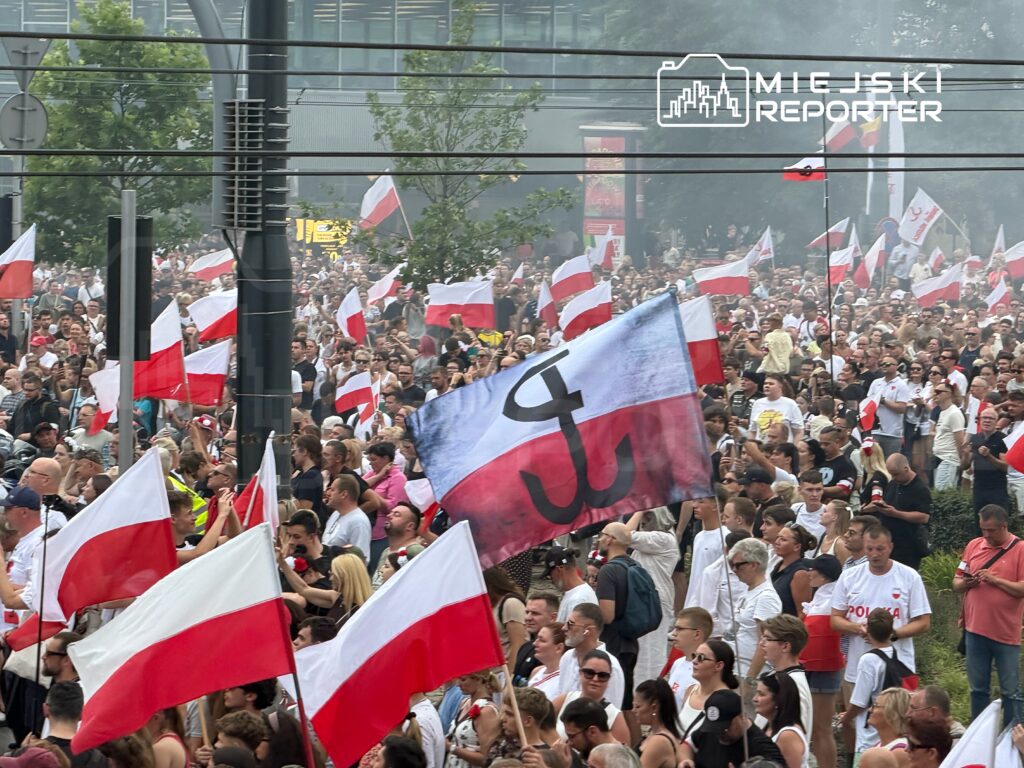 Tłum ludzi trzymających flagi, w tym jedną z symbolem Polski Walczącej, podczas manifestacji w miejskim otoczeniu.