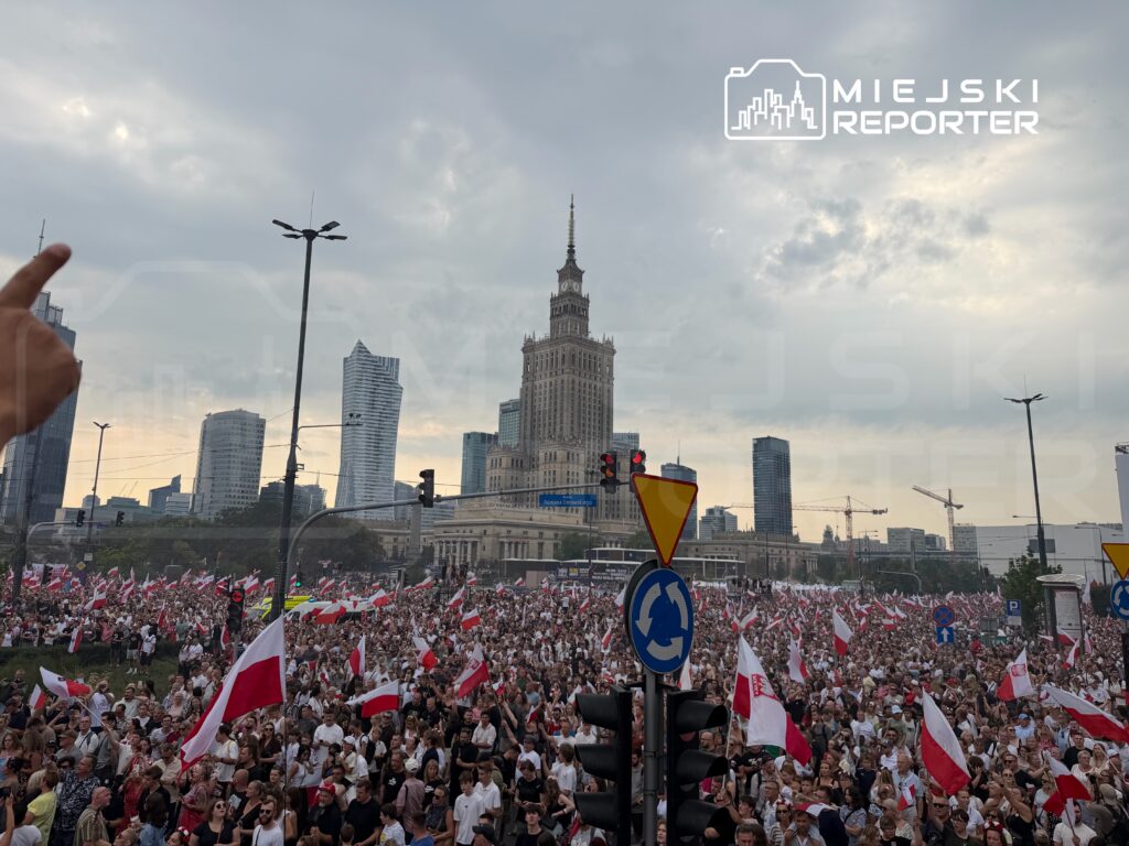Tłum ludzi z biało-czerwonymi flagami zgromadzony na ulicach w otoczeniu nowoczesnych budynków.