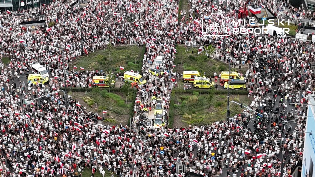 Tłum ludzi w białych i czerwonych ubraniach uczestniczy w demonstracji, otaczając karetki na ulicy.