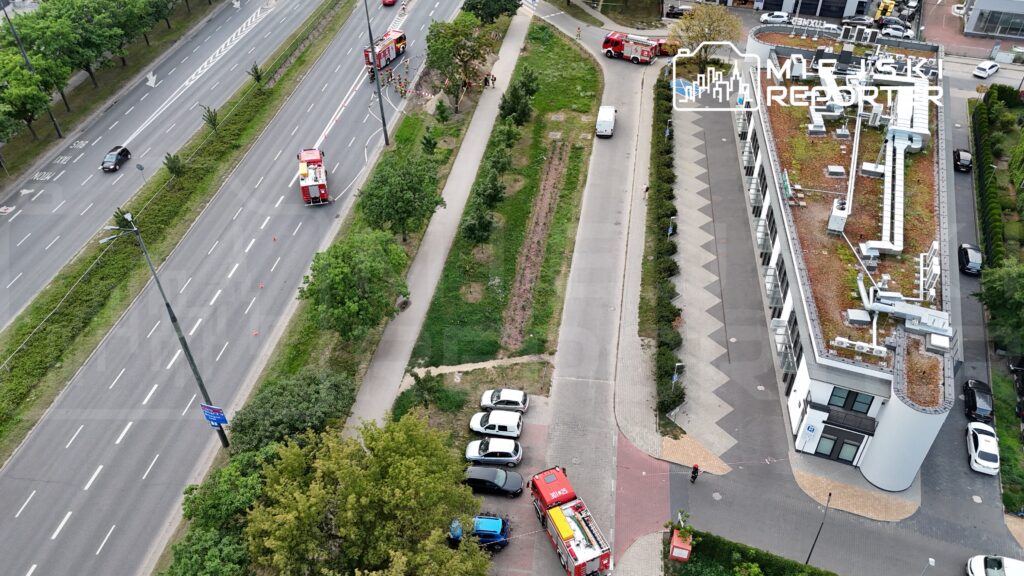 Na poboczu drogi parkują samochody, obok stoi wóz strażacki, a w tle widać ruch uliczny oraz zieleń.