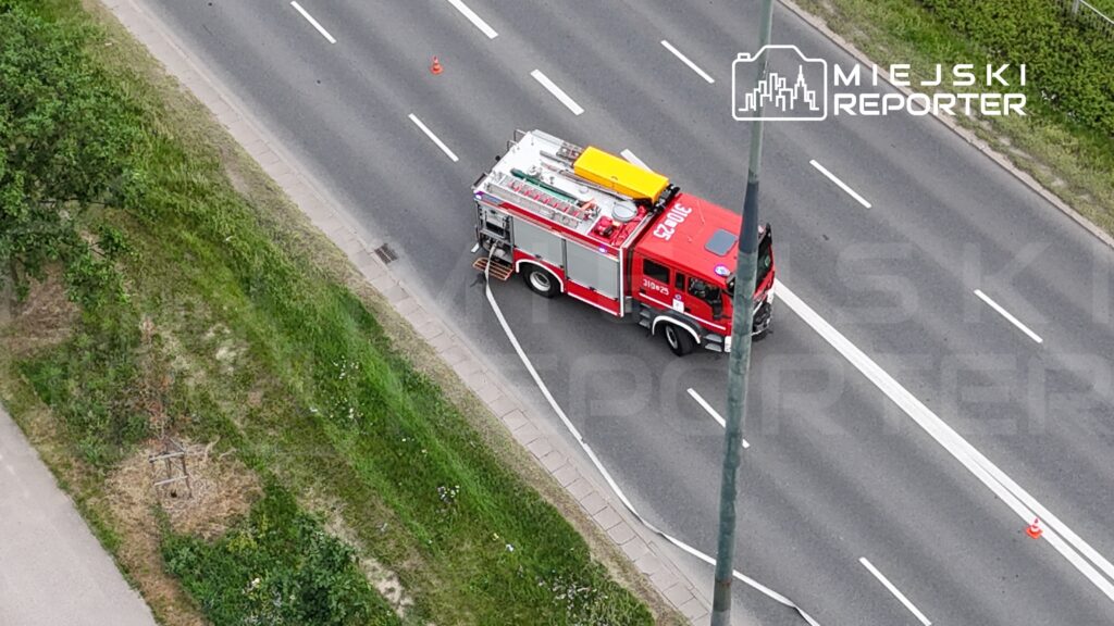 Czerwony wóz strażacki zatrzymany na poboczu drogi, z rozwiniętym wężem przeciwpożarowym.