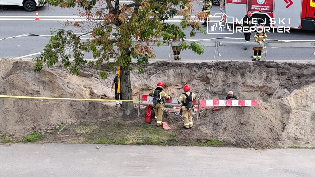 Grupa strażaków w hełmach pracuje przy wykopie, zabezpieczonym taśmą i barierkami, obok ruchliwej drogi.