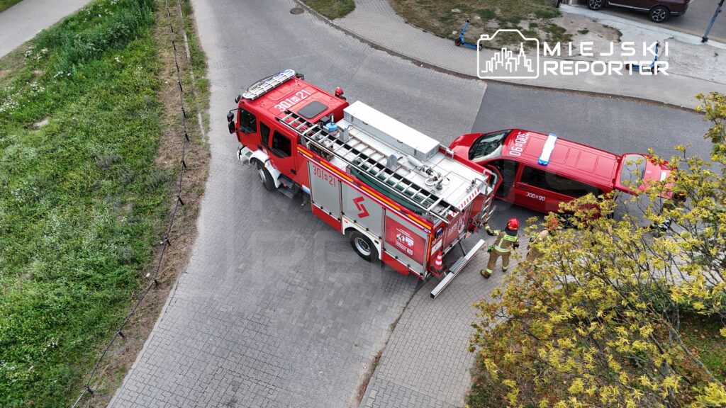 Czerwony wóz strażacki oraz pojazd ratunkowy zatrzymane na skrzyżowaniu, obok strażacy w mundurach rozmawiają.