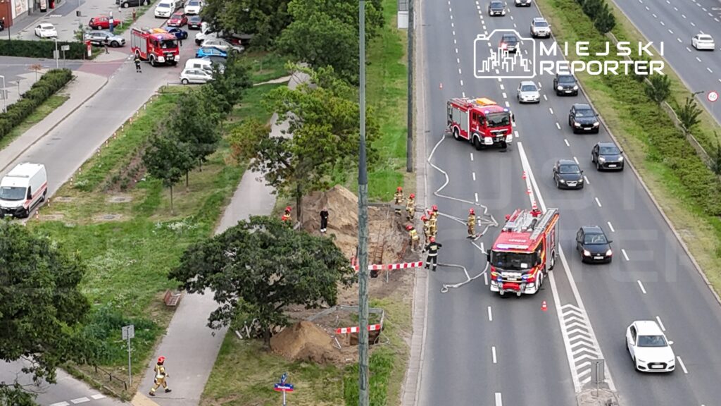 Strażacy w hełmach pracują przy wykopie na poboczu drogi, obok wozu strażackiego i zaparkowanych samochodów.