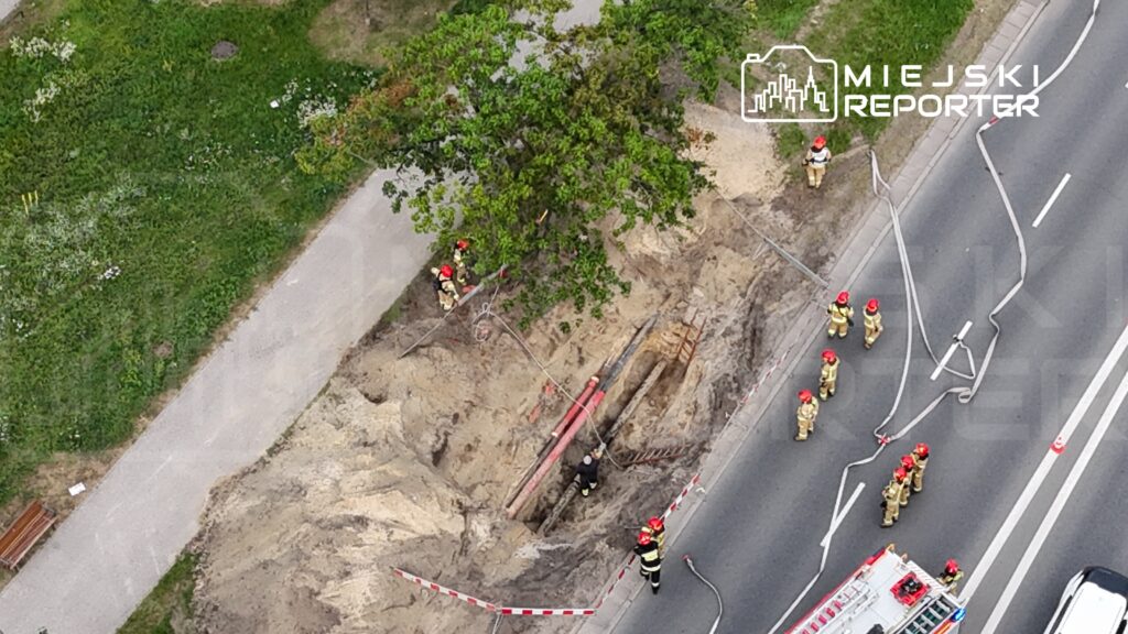 Grupa strażaków w hełmach pracuje przy wykopie drogowym, obok widocznych rur, w miejskim otoczeniu.