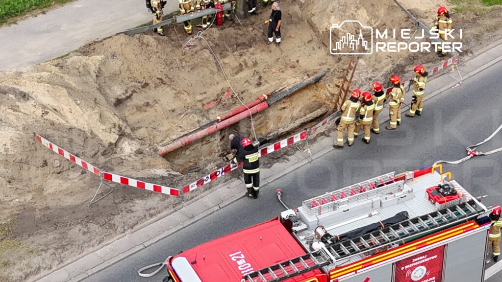 Grupa strażaków w hełmach pracuje przy wykopie drogowym, gdzie widoczne są rury i znaki ostrzegawcze.